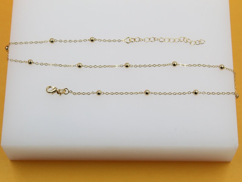 A delicate gold chain anklet featuring evenly spaced round beads rests on a white display surface.
