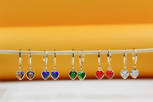 Five pairs of gold hoop earrings with colorful heart-shaped cubic zirconia charms hang in a row against an orange and white background.