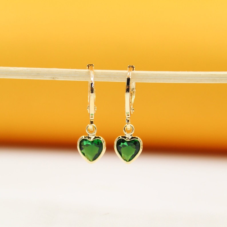 A pair of gold huggie hoop earrings featuring sparkling green heart-shaped crystal charms rests on a thin wooden stick against a warm yellow and white background.
