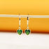 A pair of gold huggie hoop earrings featuring sparkling green heart-shaped crystal charms rests on a thin wooden stick against a warm yellow and white background.