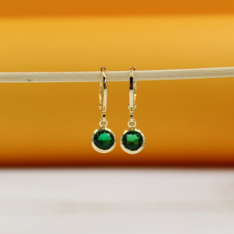 A pair of gold huggie hoop earrings featuring brilliant green gemstone drops hangs delicately from a light-colored stick against a warm orange and white background.