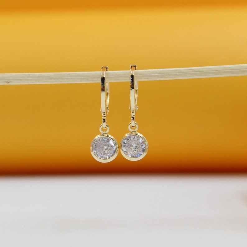 A pair of gold huggie hoop earrings featuring sparkling round cubic zirconia stone pendants hangs from a thin white stick against a warm yellow and white background.