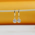A pair of gold huggie hoop earrings featuring sparkling round cubic zirconia stone pendants hangs from a thin white stick against a warm yellow and white background.