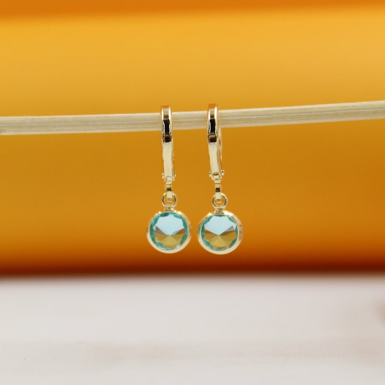 A pair of gold dangle earrings featuring sparkling light blue crystals hangs delicately on a wooden stick against a warm orange background.