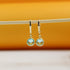 A pair of gold dangle earrings featuring sparkling light blue crystals hangs delicately on a wooden stick against a warm orange background.