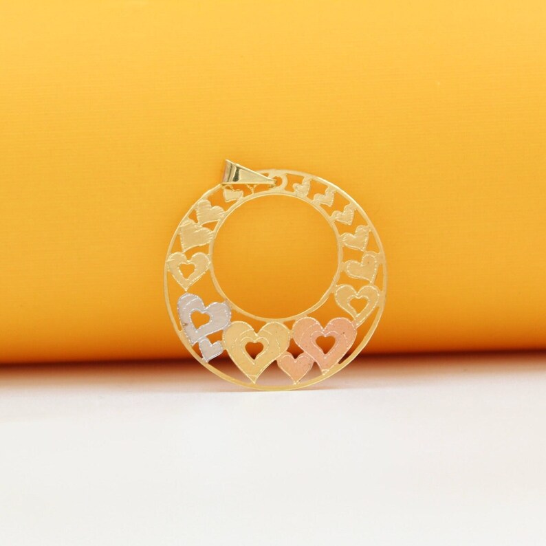 A tri-color gold pendant features an intricate circular design filled with interlocking cut-out hearts against a solid yellow backdrop.