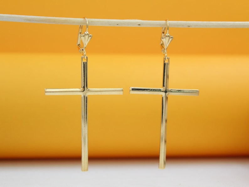 A pair of elegant gold cross dangle earrings hangs suspended against a bright yellow background.