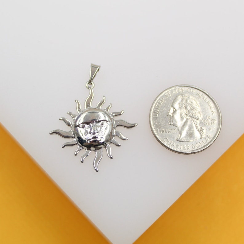 A silver sun face pendant rests next to a US quarter coin for scale on a white and orange surface.