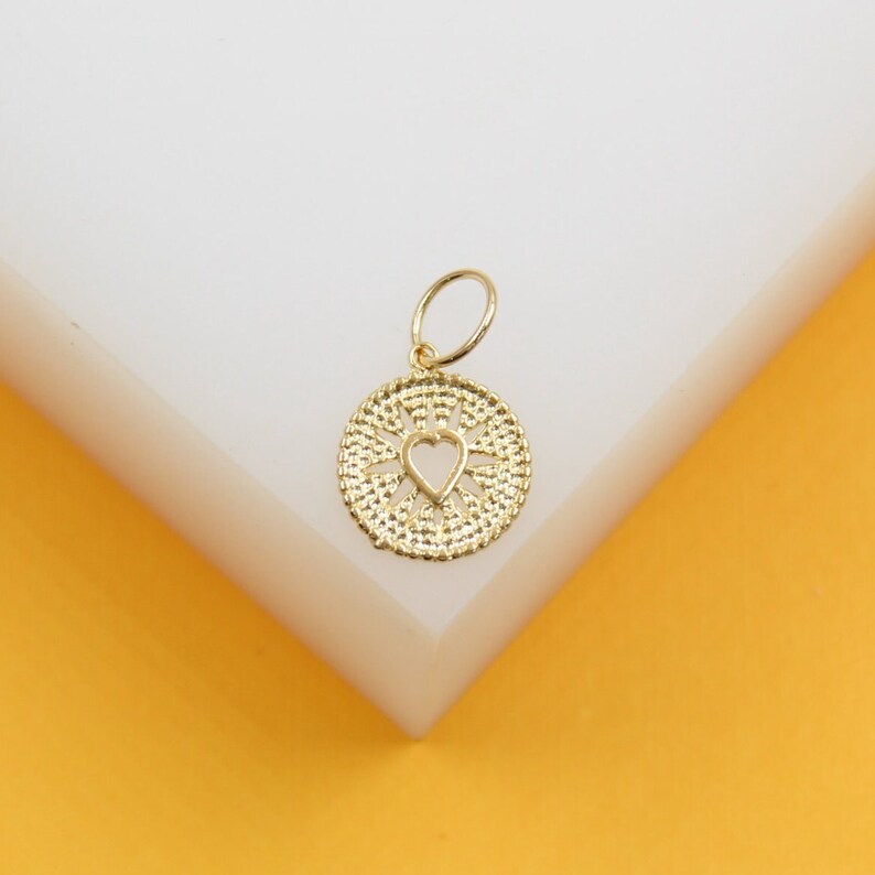 A gold-plated charm rests on a white display block, featuring an intricate sunburst pattern and a central cutout heart.