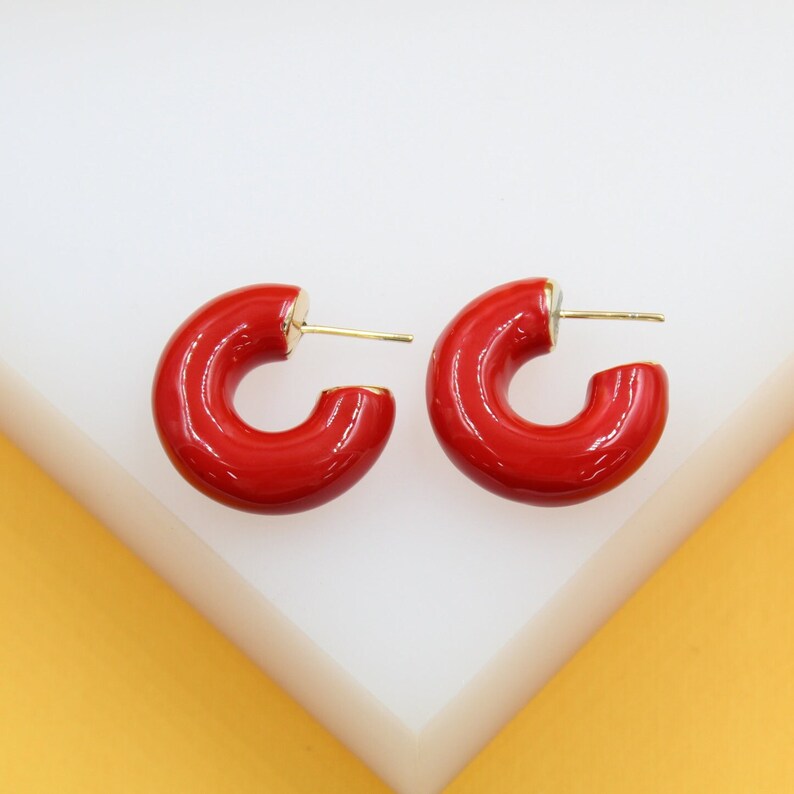 A pair of glossy red enamel hoop earrings rests on a white display stand.
