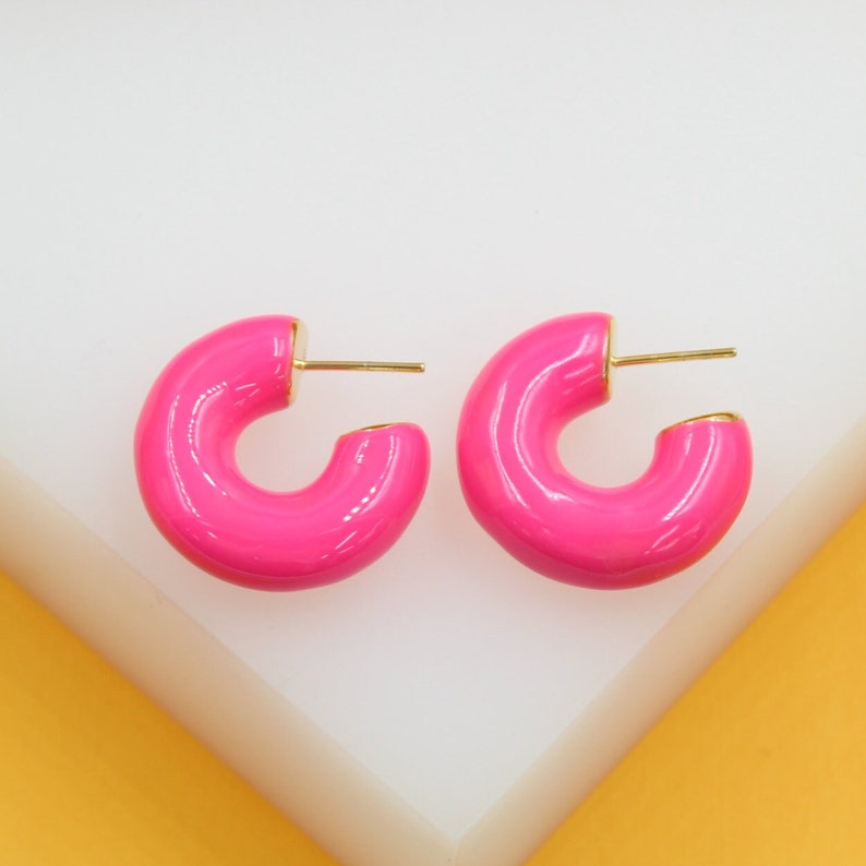 A pair of glossy hot pink chunky hoop earrings with gold-plated posts rest on a white display surface.