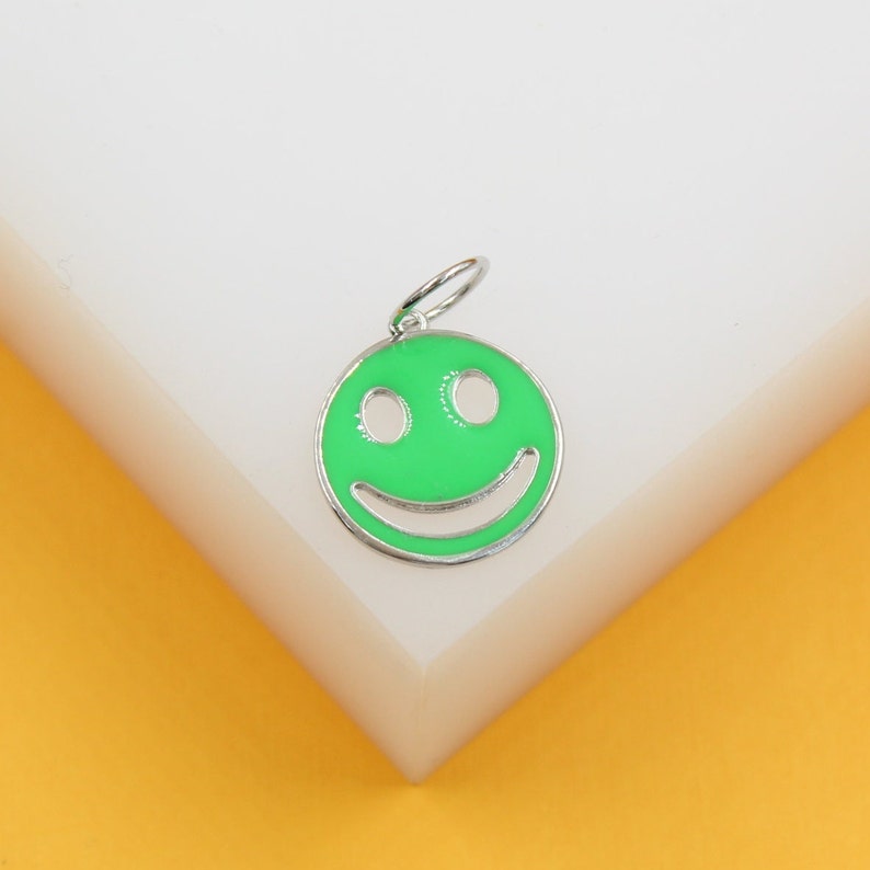 A silver smiley face charm with bright green enamel rests on a white display block against a yellow background.