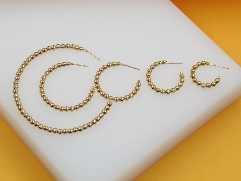 Five gold beaded hoop earrings in graduated sizes rest on a white display surface against an orange background.