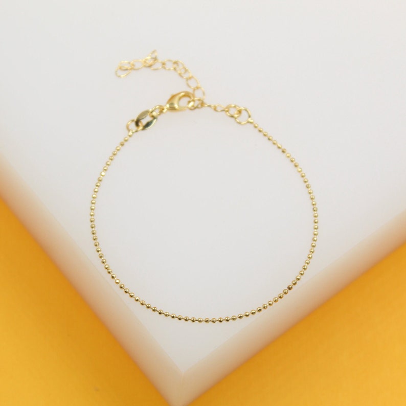 A delicate gold ball chain bracelet rests on a white display surface against a vibrant yellow background.