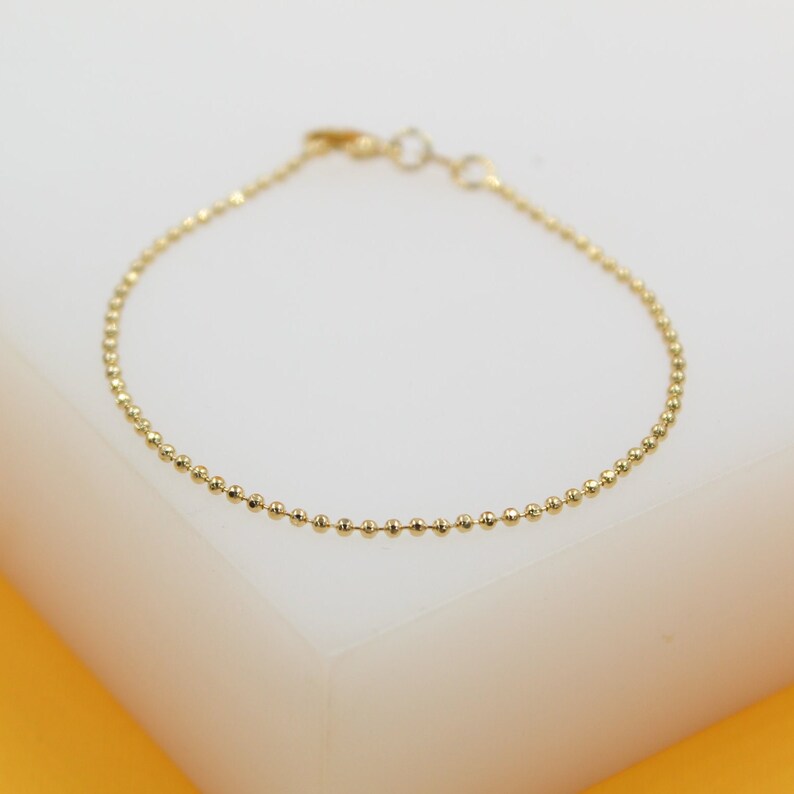 A delicate gold ball chain bracelet rests on a clean white display surface against a vibrant yellow background.
