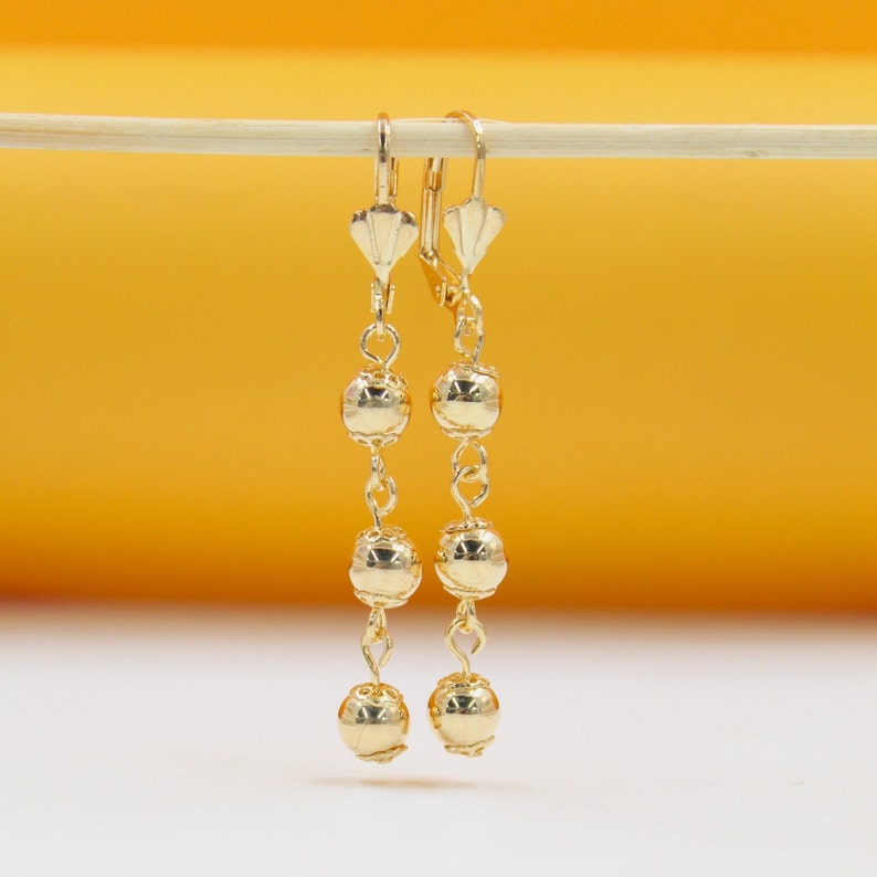 A pair of elegant gold-plated dangle earrings featuring graduated spherical beads hang gracefully against a warm yellow and white background.