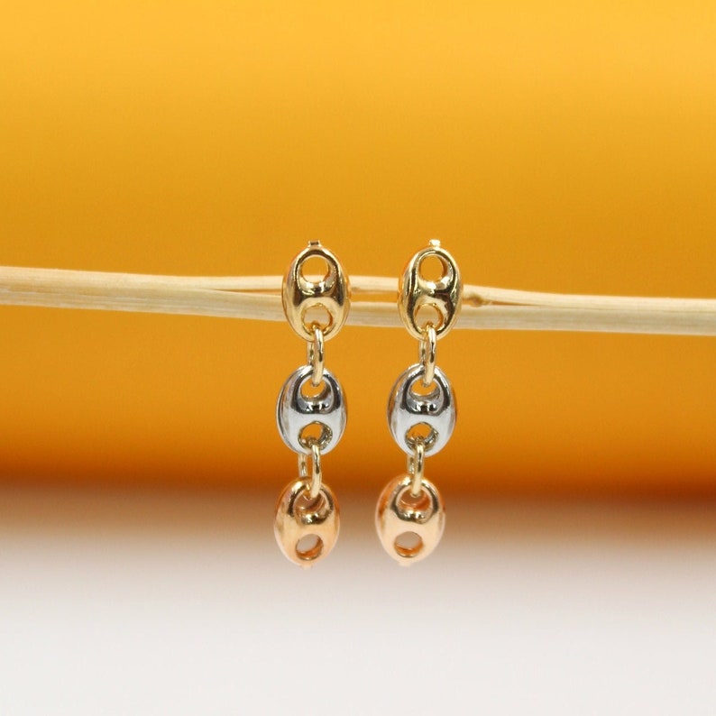 A pair of tri-color gold dangle earrings featuring interlocking mariner links hangs delicately from a wooden stick against a warm yellow and white background.