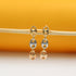 A pair of tri-color gold dangle earrings featuring interlocking mariner links hangs delicately from a wooden stick against a warm yellow and white background.