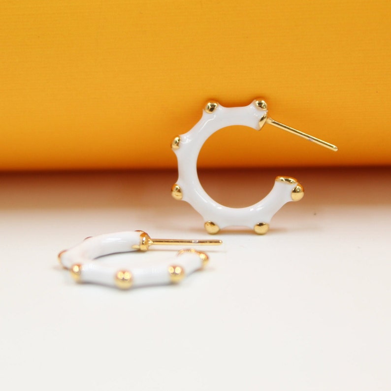 A pair of white enamel hoop earrings featuring gold bead accents rests against a bright yellow and white background.