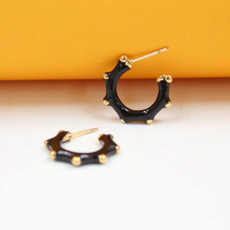 Two black enamel and gold-plated hoop earrings with a bumpy texture rest on a white surface against an orange background.