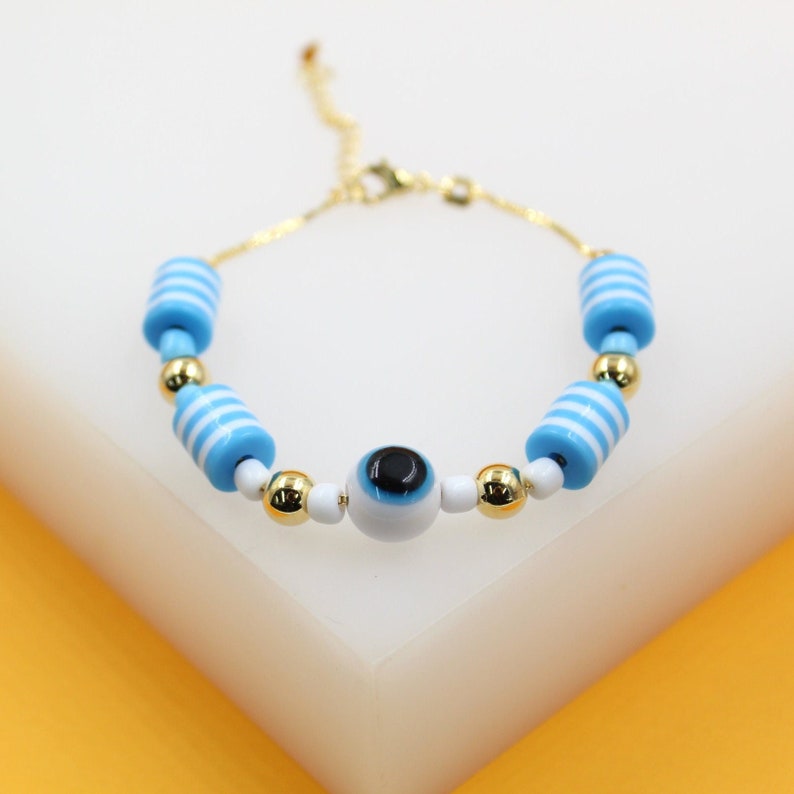 A delicate gold chain bracelet features blue and white striped cylindrical beads alongside a central evil eye charm on a white surface.