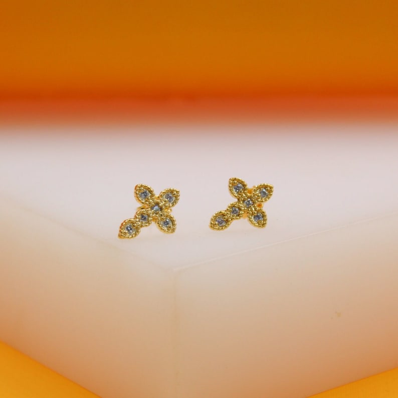 A pair of gold cross stud earrings adorned with sparkling cubic zirconia stones rests upright on a light display surface.