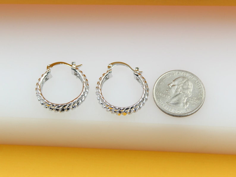 A pair of textured silver hoop earrings sits next to a US quarter coin on a white surface to show their scale.