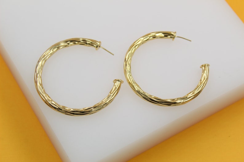 A pair of textured gold hoop earrings rests on a white display block against an orange background.