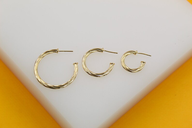 Three graduated sizes of twisted gold hoop earrings rest neatly on a white display surface against a vibrant yellow background.