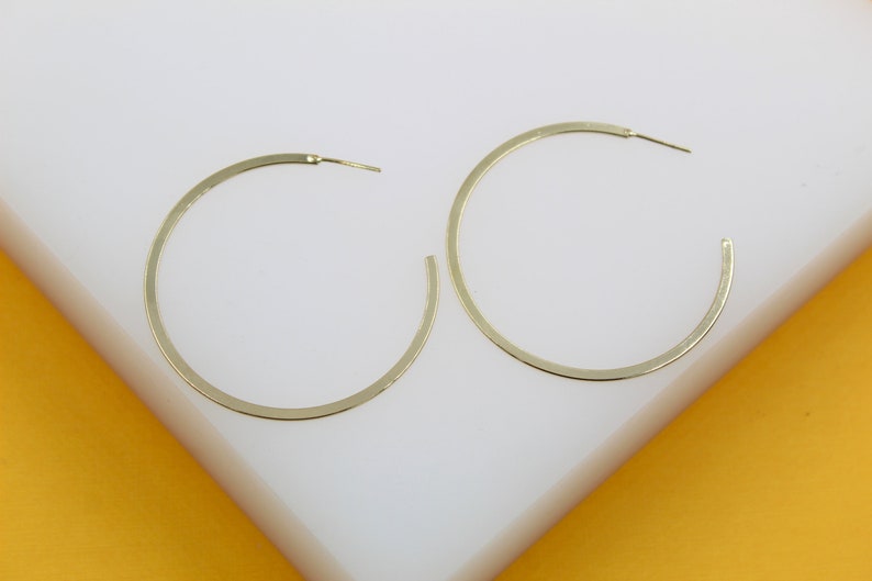 Two large gold hoop earrings rest neatly on a white display surface against a vibrant yellow background.