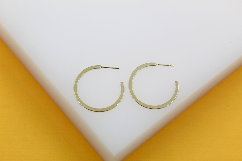 A pair of minimalist flat gold hoop earrings rests on a white and yellow display stand.