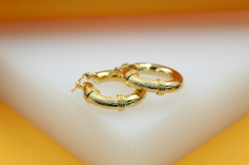 Two textured gold hoop earrings rest on a white surface against an orange background.