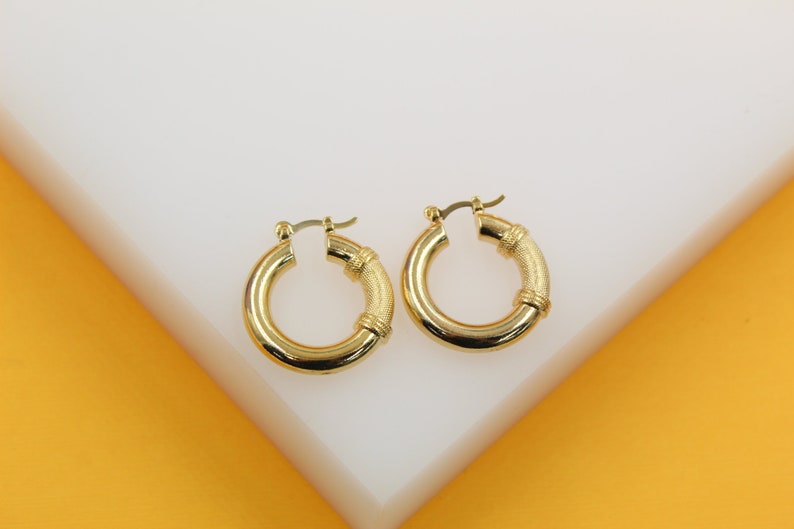 A pair of thick gold hoop earrings rests on a white display surface against a vibrant yellow background.