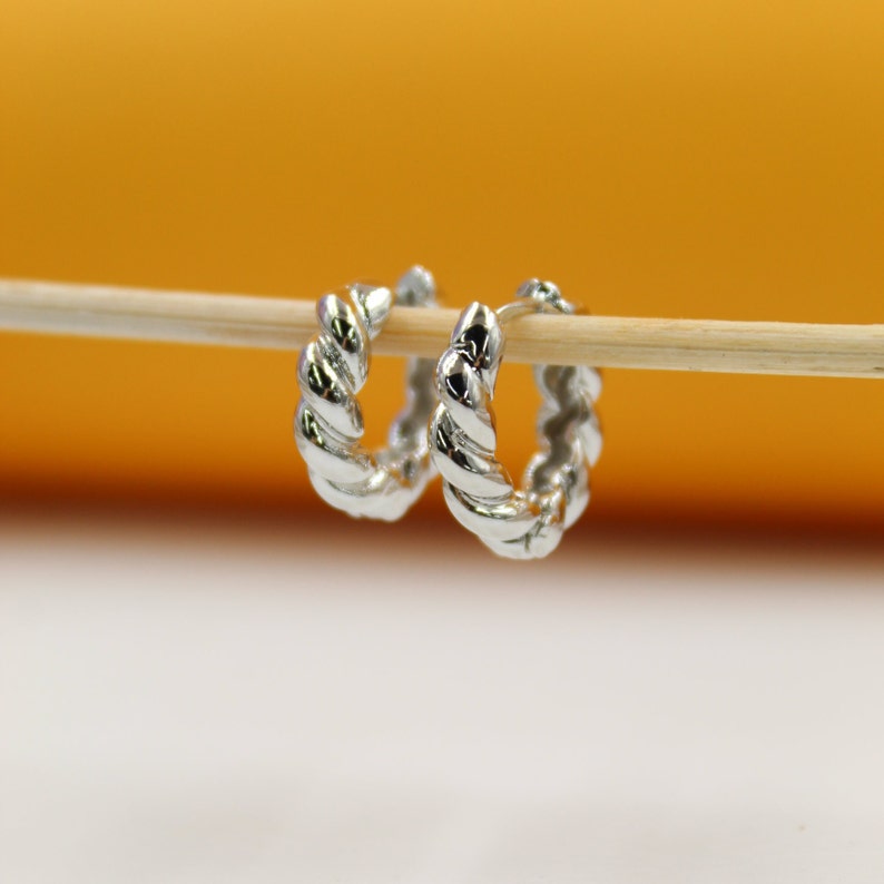 A pair of polished silver twisted hoop earrings hangs neatly from a wooden stick against a warm, blurred background.