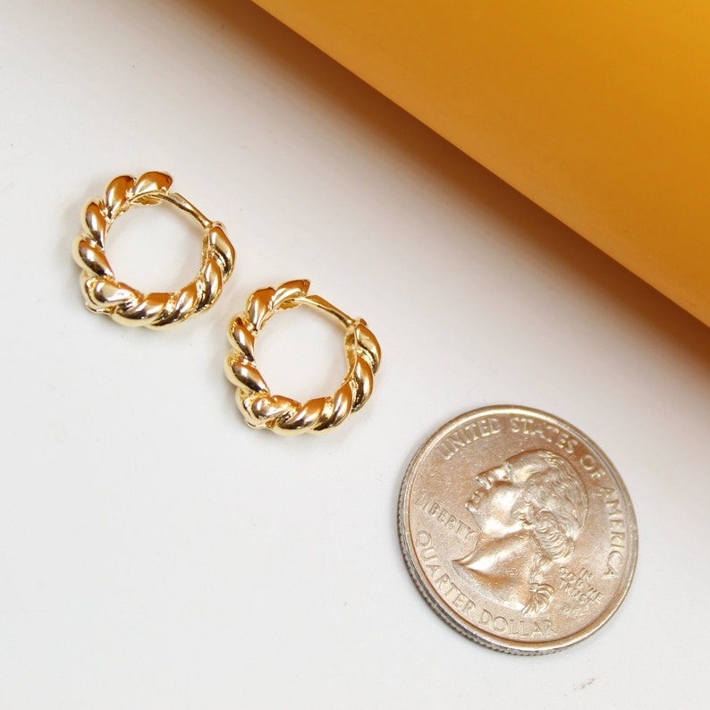 A pair of polished gold twist hoop earrings rests next to a US quarter coin for scale on a white surface.