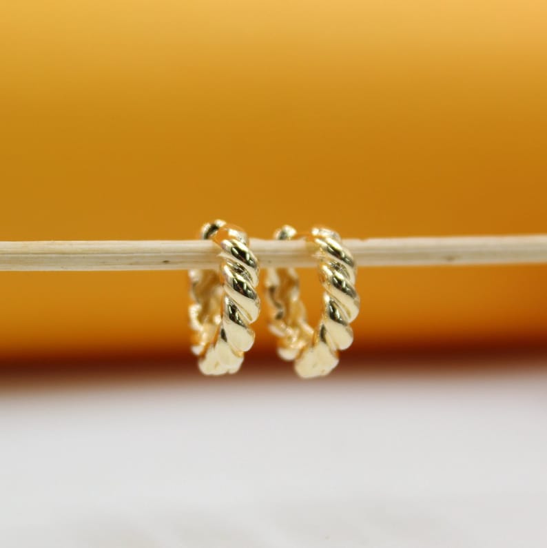 A pair of shiny gold twisted huggie hoop earrings hang delicately from a wooden stick against a warm yellow and white background.