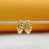A pair of shiny gold twisted huggie hoop earrings hang delicately from a wooden stick against a warm yellow and white background.
