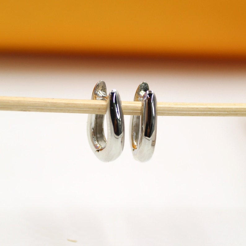 A pair of polished silver hoop earrings hangs delicately from a wooden stick against a soft-focus background.