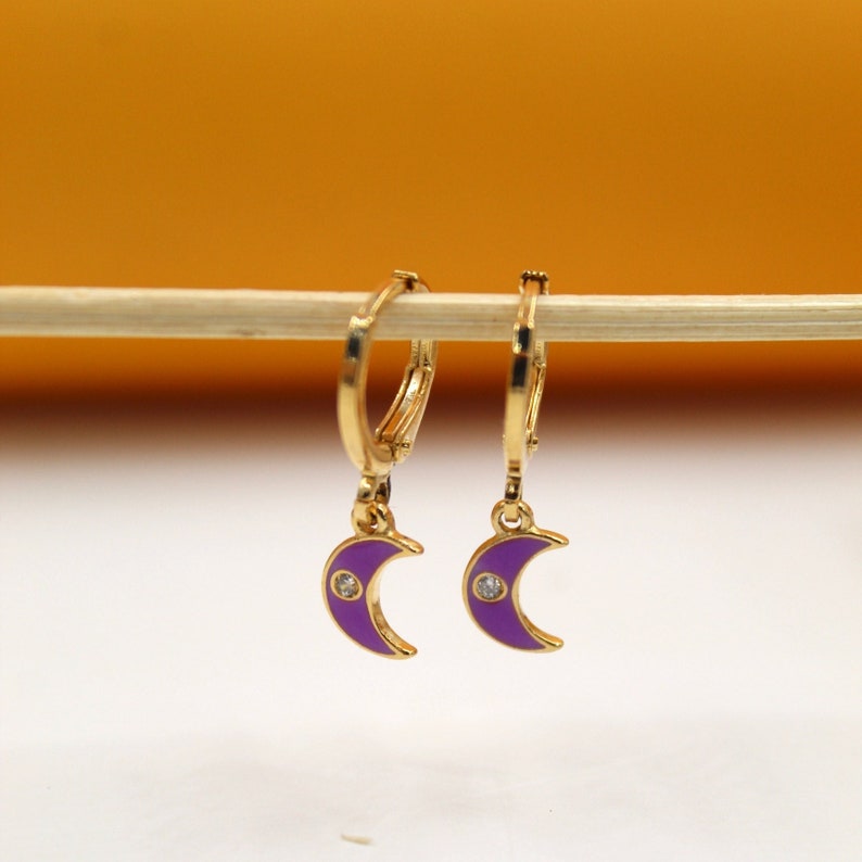 A pair of gold hoop earrings with purple enamel crescent moon charms hang delicately from a wooden stick.