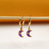 A pair of gold hoop earrings with purple enamel crescent moon charms hang delicately from a wooden stick.