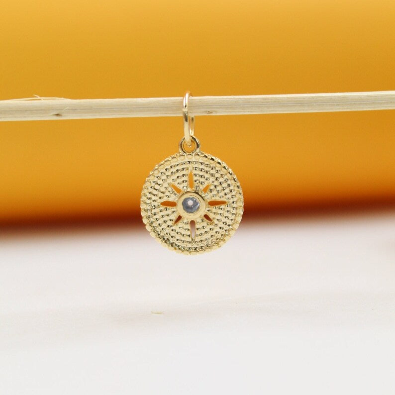 A gold sun-burst charm hangs delicately from a horizontal stick against a warm, neutral background.