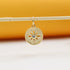A gold sun-burst charm hangs delicately from a horizontal stick against a warm, neutral background.