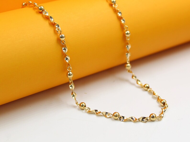 A delicate gold chain necklace with alternating smooth round beads and twisted links rests gracefully against a bright orange and white background.