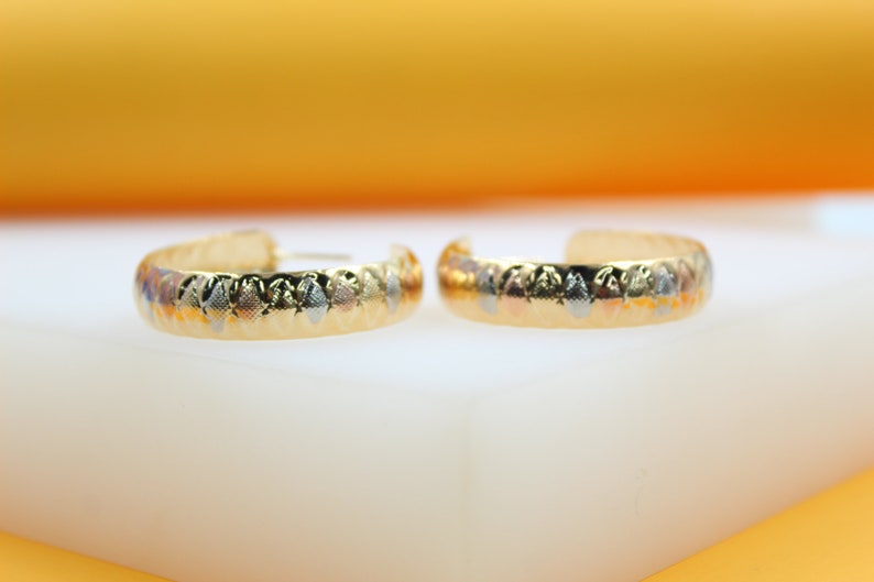 A pair of textured gold hoop earrings with alternating silver and gold diamond-shaped patterns rests on a white surface against an orange background.