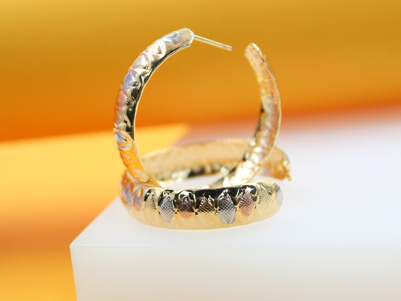 A pair of elegant tri-color gold hoop earrings rests on a white display surface against a warm yellow background.