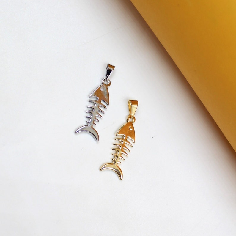 A silver fish skeleton pendant and a gold fish skeleton pendant rest side-by-side on a white surface next to a yellow background.