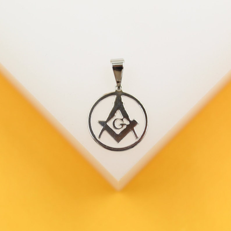 A polished silver Masonic square and compasses pendant rests on a white display against a vibrant yellow background.