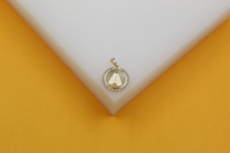 A gold initial A pendant rests on a white block against a warm yellow background.