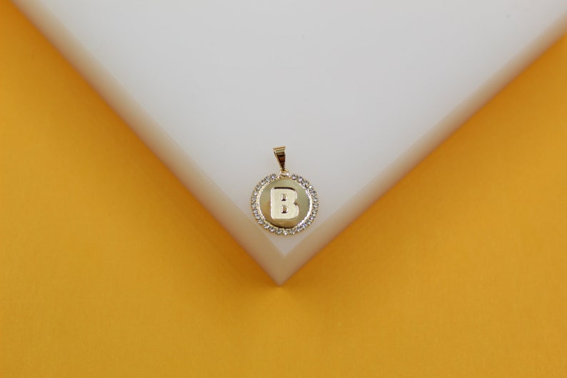 A gold initial B pendant necklace sparkles with a halo of small diamonds against a white and yellow background.