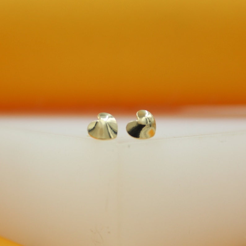 A pair of shiny gold heart stud earrings rests on a white display surface against a warm yellow background.